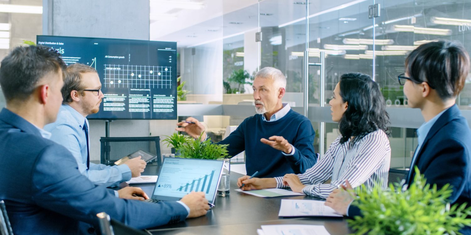 Senior Executive Explains Company's Vision and Potential to His Employees. They are Sitting at Big Table in Meeting Room. TV Screen on the Wall Shows Corporate Growth.