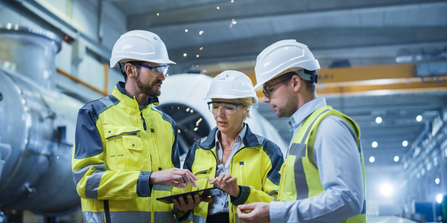 Three Heavy Industry Engineers Stand in Pipe Manufacturing Factory, Use Digital Tablet Computer, Have Discussion. Large Pipe Assembly. Design and Construction of Oil, Gas and Fuels Transport Pipeline