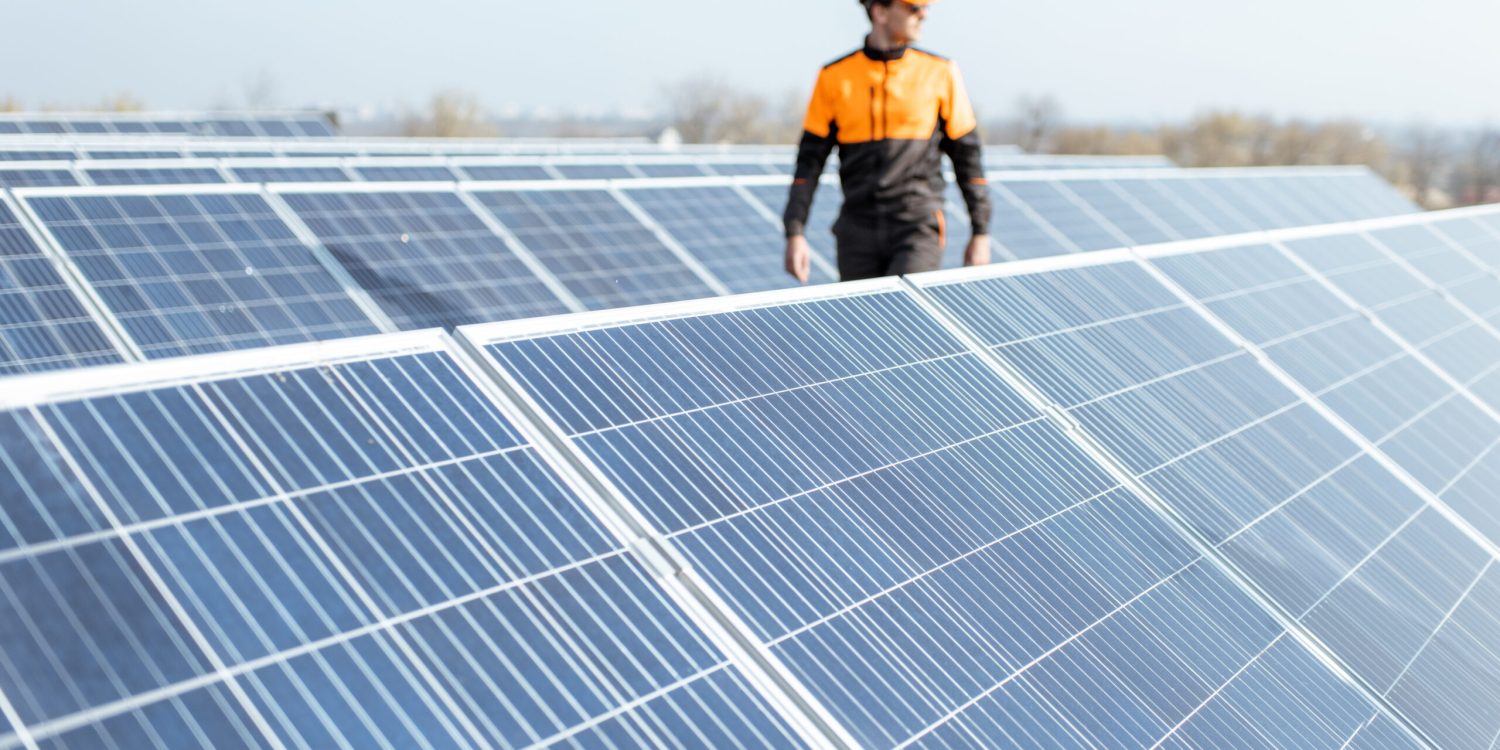 View on the rooftop solar power plant with engineer in protective workwear walking and examining photovoltaic panels. Concept of alternative energy and its maintenance