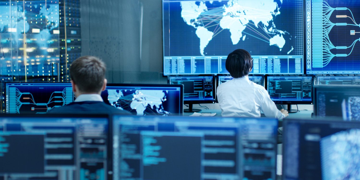 In the System Control Room Operator and Administrator Sitting at Their Workstations with Multiple Displays Showing Graphics and Logistics Information.